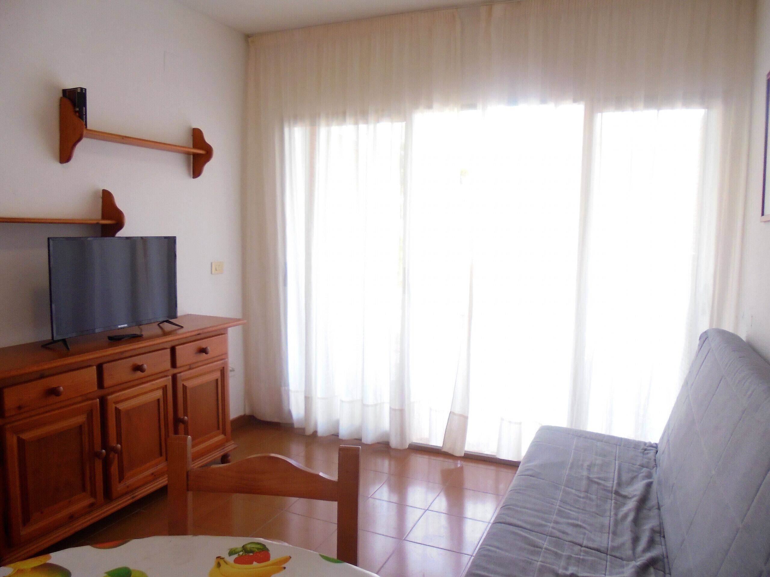Ein Zimmer mit einem Holzschrank, einem Fernseher darauf, einem grauen Sofa und einem großen Fenster mit durchsichtigen Vorhängen.