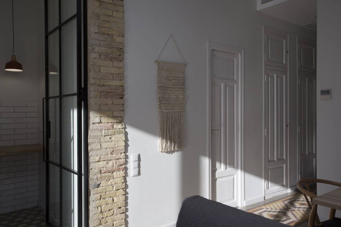 Une pièce avec un mur en briques, des portes blanches, une décoration murale en macramé et une chaise en bois.