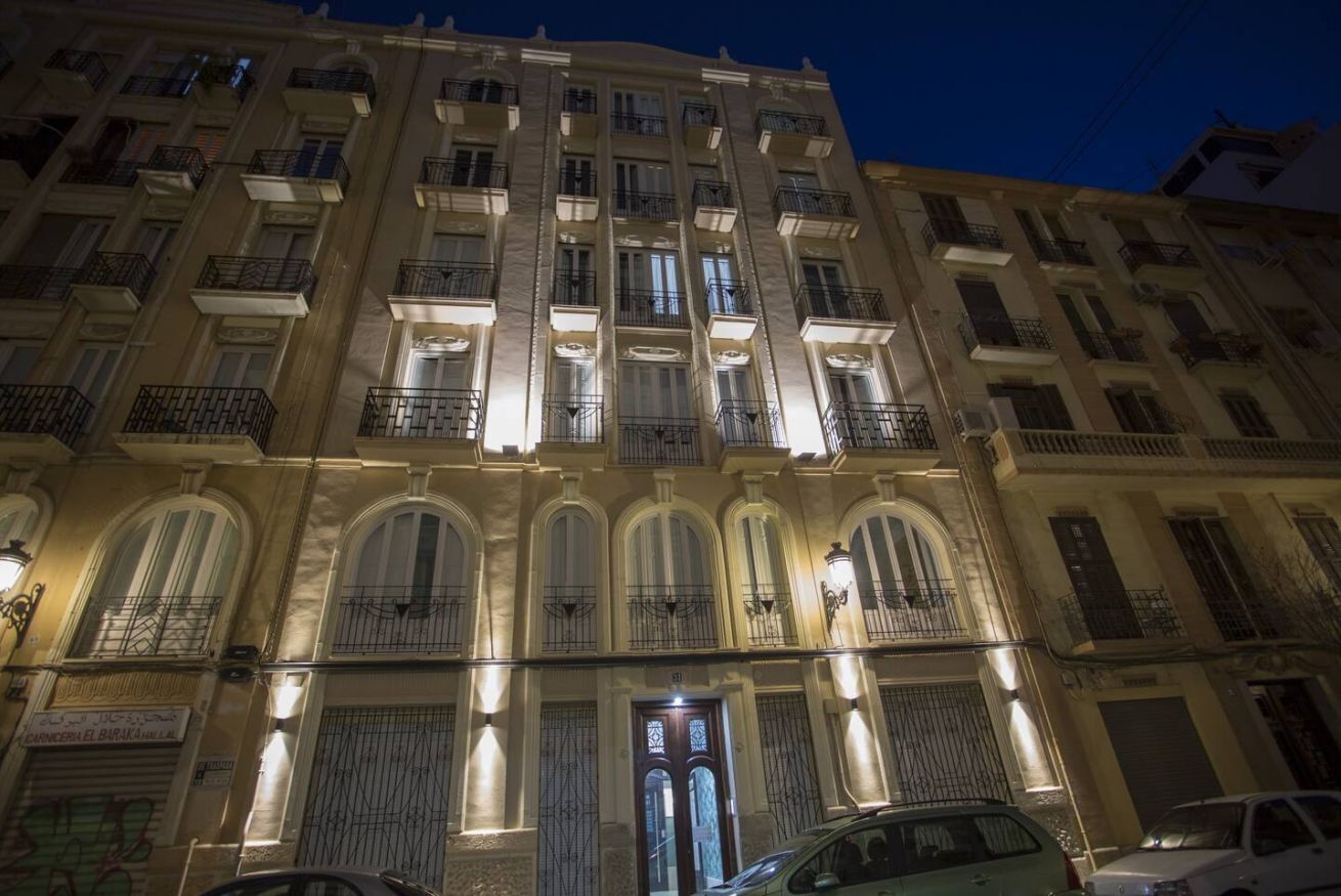 Un immeuble à plusieurs étages, doté de balcons et de fenêtres en arc de cercle, avec une entrée centrale surmontée d’une porte et d’une enseigne.