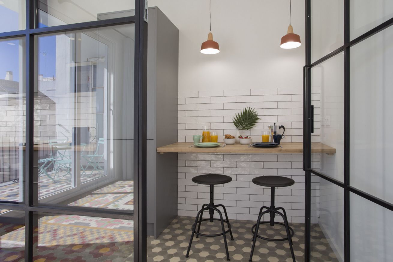 Une cuisine moderne avec une porte vitrée donnant sur un balcon, deux tabourets et des luminaires suspendus.