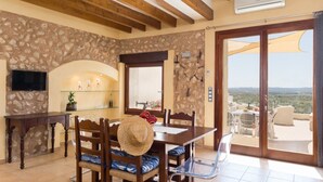A dining area with a stone wall, wooden ceiling beams, a TV, and a view of the landscape through a glass door. - Casa del Sol - Beautiful town house with pool and views of the countryside (Búger)
