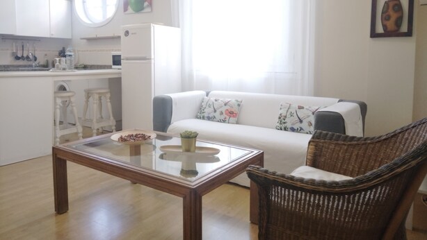 Ein modernes Wohnzimmer mit Glastisch, einem weißen Sofa, einem Korbsessel, einer Küchenzeile mit weißen Schränken und einem großen Fenster mit Vorhängen.