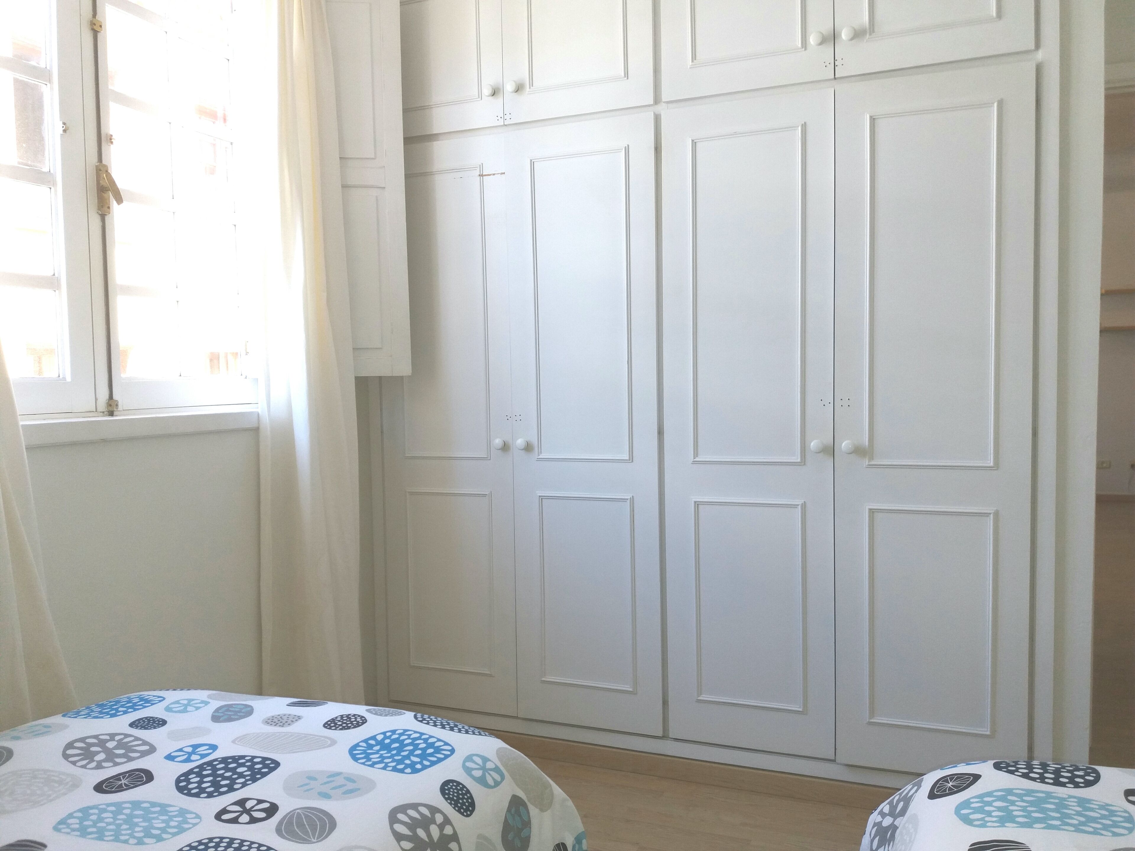 A room with a large white wardrobe, a bed with a patterned bedspread, and a window with curtains.