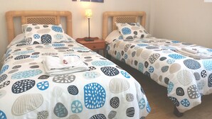 Ein Doppelzimmer mit zwei Betten, jeweils mit gestecktem Bezug und Kissen, einem Nachttisch mit Lampe sowie einem Wandbild über den Betten.