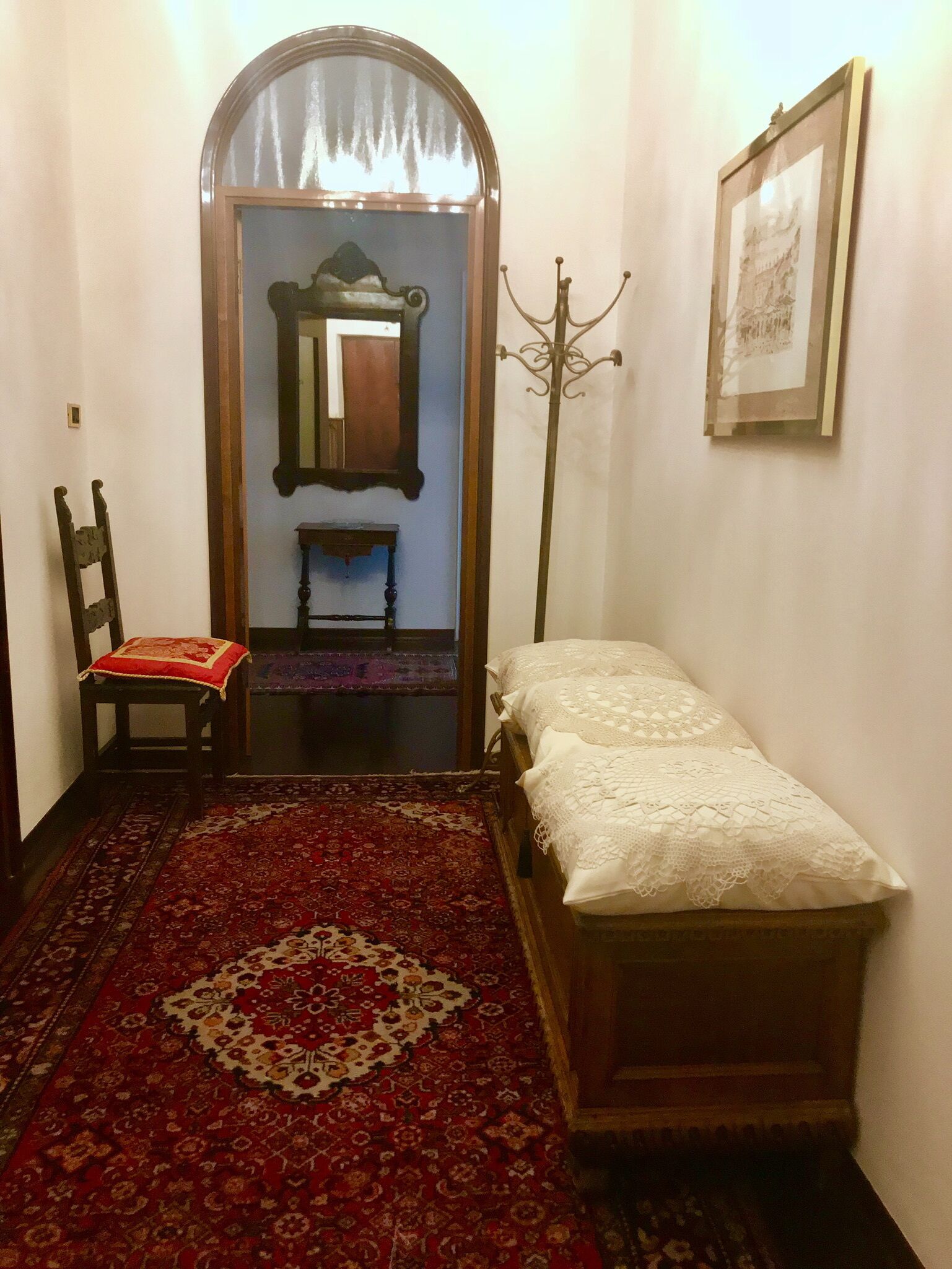A narrow room with a wooden bed, a red patterned rug, a wooden chair, a small table, a mirror, and a framed picture on the wall.