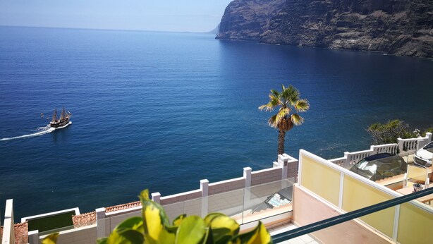 A coastal view with a boat on the water, a palm tree, and a building with a balcony overlooking the sea.