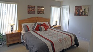 Une chambre à coucher avec un grand lit, deux tables de chevet, une petite table, une plante en pot et des œuvres d’art accrochées aux murs.