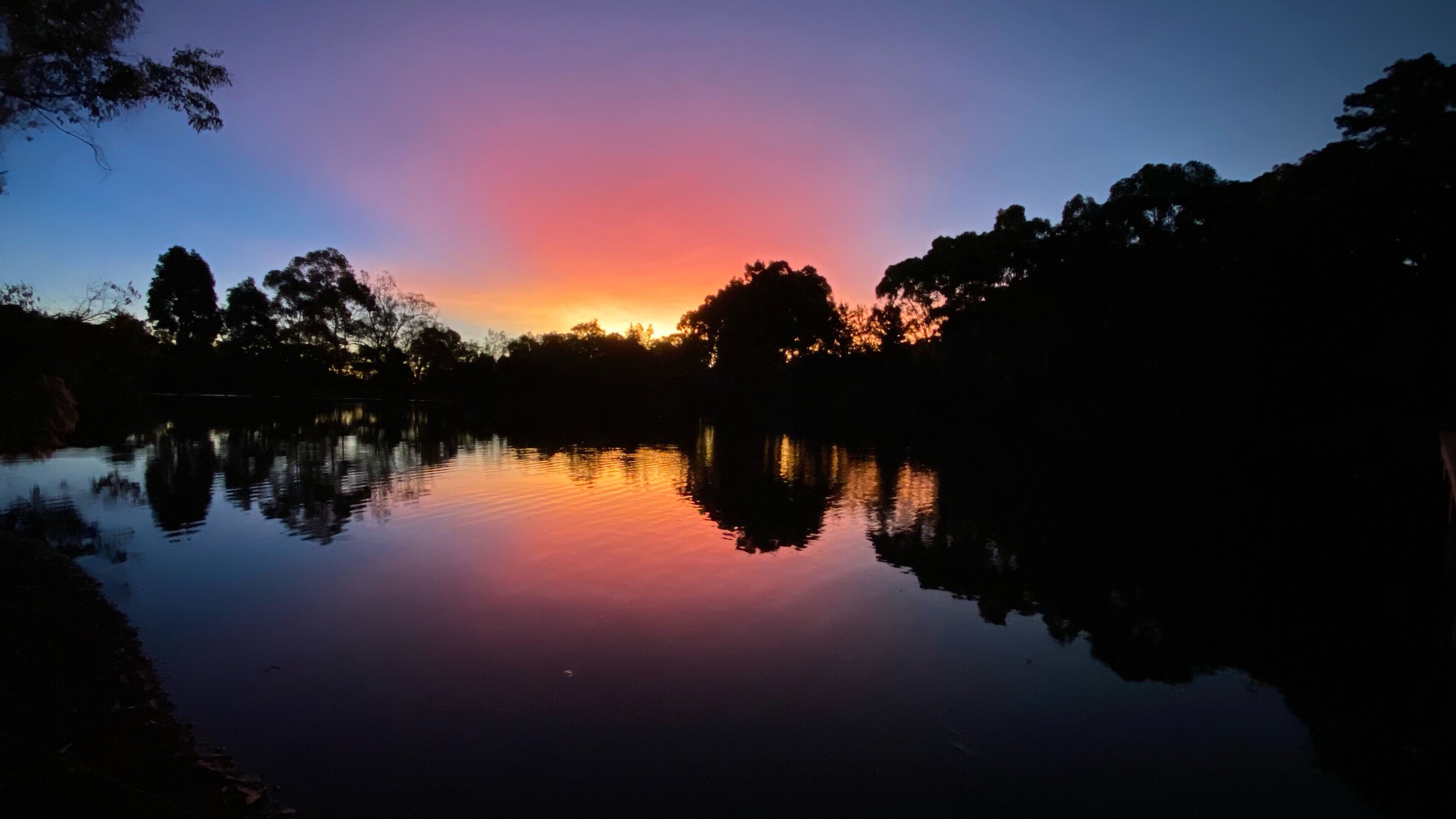 Un llac tranquil que reflecteix el capvespre, amb siluetes d'arbres i un cel clar.