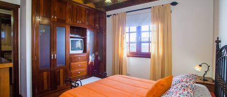 A bedroom with a wooden wardrobe, a bed with an orange bedspread, a patterned pillow, and a window with curtains.