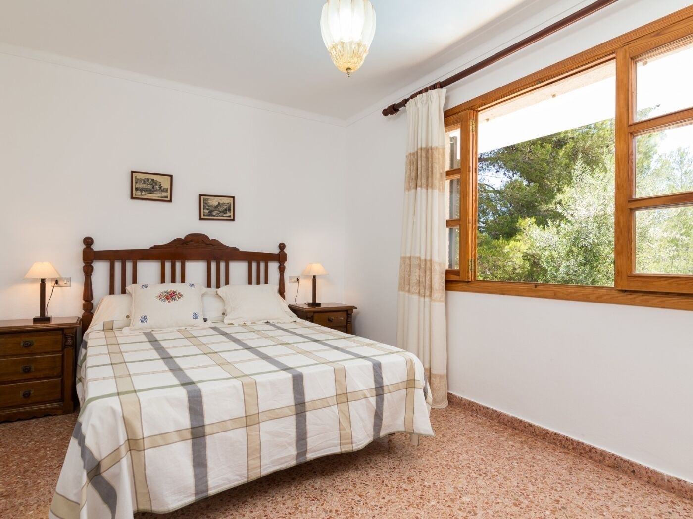 A bedroom with a bed, two bedside tables, a window with curtains, and two wall-mounted pictures.