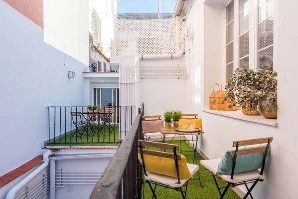 Une petite cour étroite avec une table et des chaises, une fenêtre ornée de plantes et une porte.