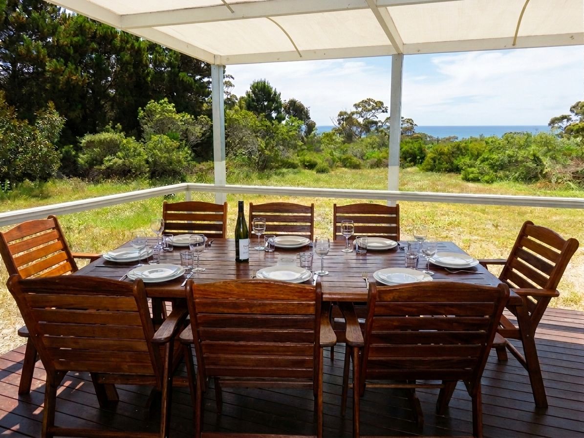 An outdoor dining area with a table set for six, surrounded by wooden chairs, under a covered structure with a view of the landscape.