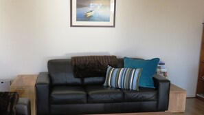 A black leather sofa with two pillows, a framed picture above it, and a wooden side table.