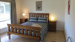 A bedroom with a wooden bed, two bedside tables with lamps, a window with curtains, and a wall-mounted artwork.