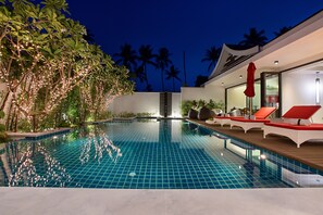 A swimming pool with a tiled deck, surrounded by palm trees and plants, and a modern building with a glass door and red lounge chairs.