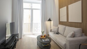 A modern living room with a sofa, a coffee table with a book and fruit, a TV, and a balcony with curtains.