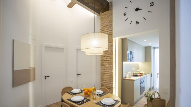 A dining area with a round table set for four, a hanging light fixture, and a kitchen visible through an open doorway.