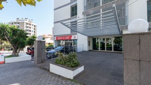 Ein modernes Gebäude mit einem 'Rent a Car'-Schild, einer überdachten Einfahrt und einem Parkplatz mit Autos.