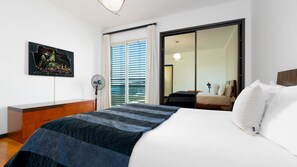 A hotel room with a bed, a desk, a chair, a fan, a painting, and a sliding door to a balcony.