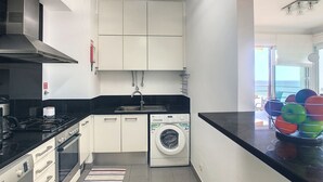 Een moderne keuken met witte kasten, een zwart aanrecht, een ingebouwde kookplaat, een wasmachine en uitzicht op zee door een groot raam.