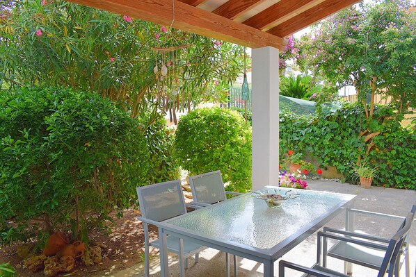 Un patio al aire libre con una mesa de vidrio y sillas de metal, rodeado de abundante follaje y plantas en macetas.