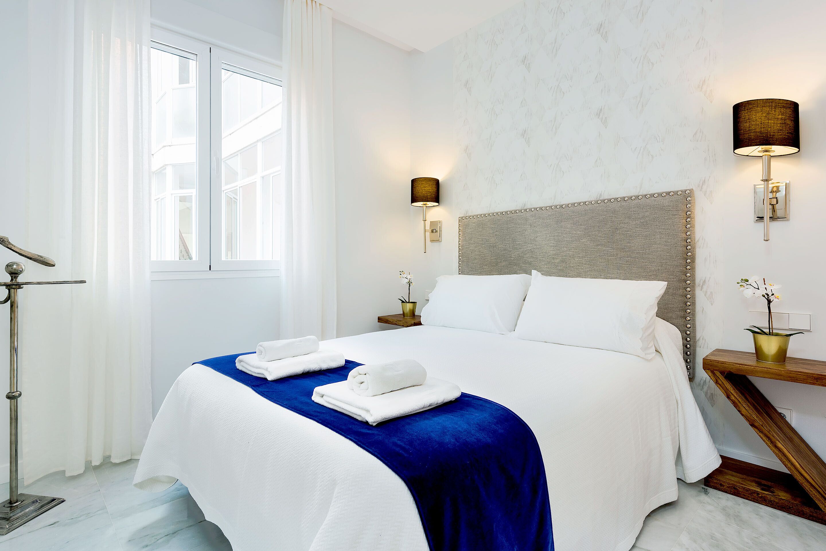 Een hotelkamer met een groot bed, twee bedlampen, een nachtkastje met een vaas en een raam met gordijnen.