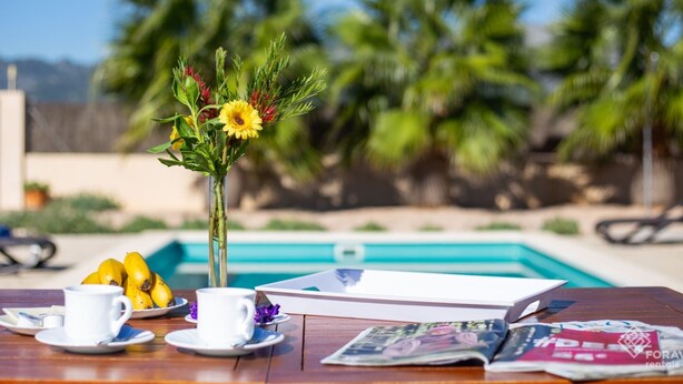 Une table en bois sur laquelle se trouvent un vase de fleurs jaunes, une assiette de bananes, deux tasses blanches et un magazine.