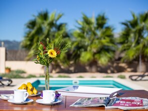 A dining table with a vase of flowers, a plate of bananas, and a cup, set against a poolside view with palm trees.