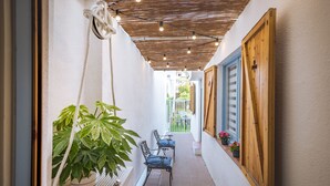 Un estrecho pasillo exterior con techo de madera, una planta colgante, dos sillas y una puerta de madera.