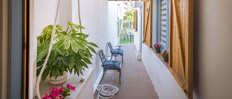 A narrow outdoor corridor with a wooden ceiling, a hanging plant, two chairs, and a wooden door.