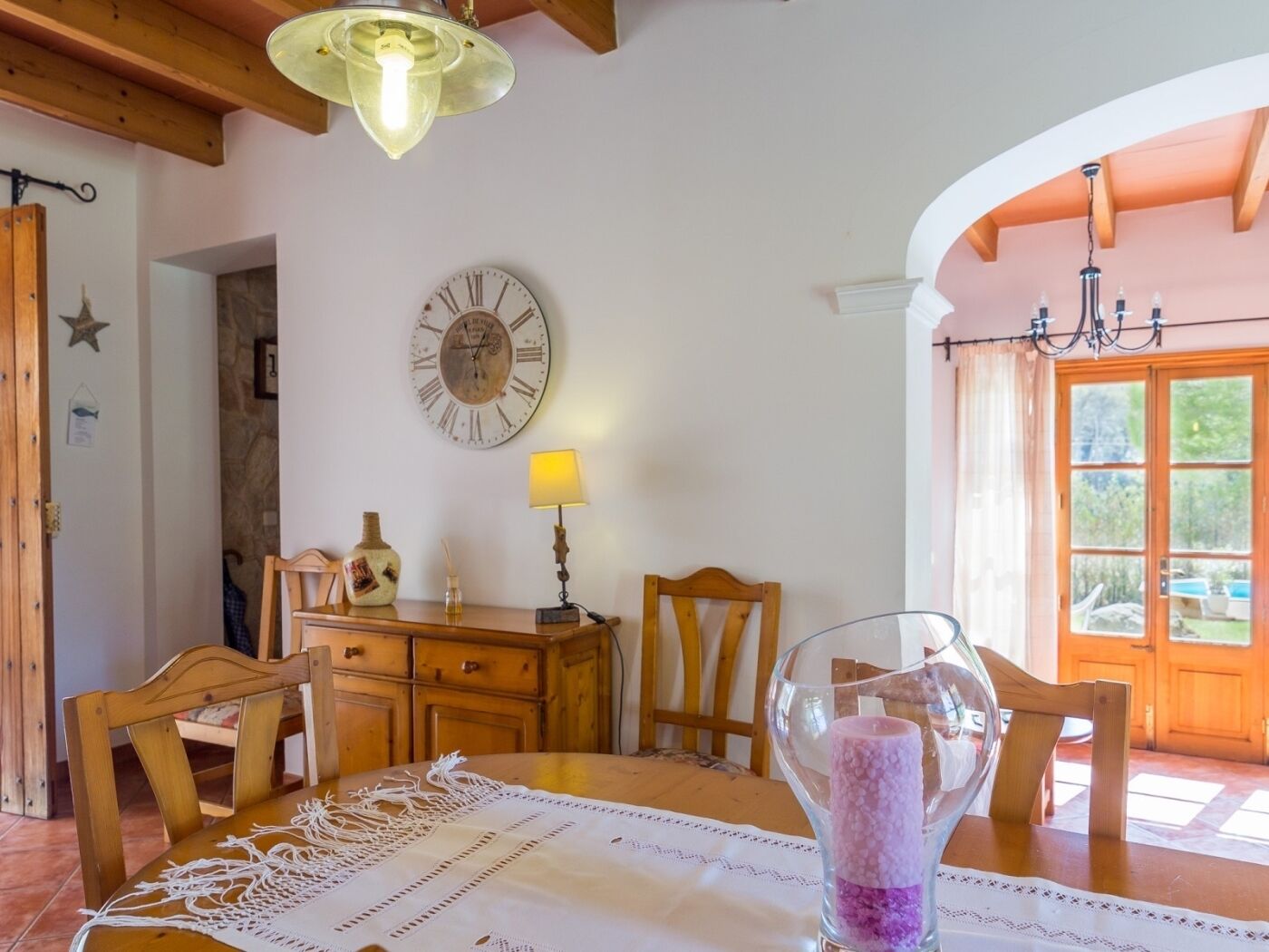 A dining area with a wooden table, chairs, a wall clock, and a window with a view of greenery.