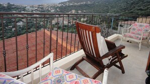 Un balcon avec vue sur la ville et les montagnes. On y trouve deux chaises en bois et une table recouverte d’une nappe colorée.