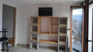 Une pièce dotée d’une bibliothèque intégrée en bois et en béton, d’un téléviseur fixé au-dessus, et d’une porte en verre donnant sur un balcon avec vue.