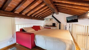 Un dormitorio con techo inclinado, una cama con ropa de cama roja y beige, una silla roja, un televisor montado en la pared y un pequeño calentador.