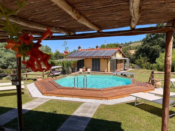 Una piscina con muelle de madera, un porche cubierto y una casa al fondo.