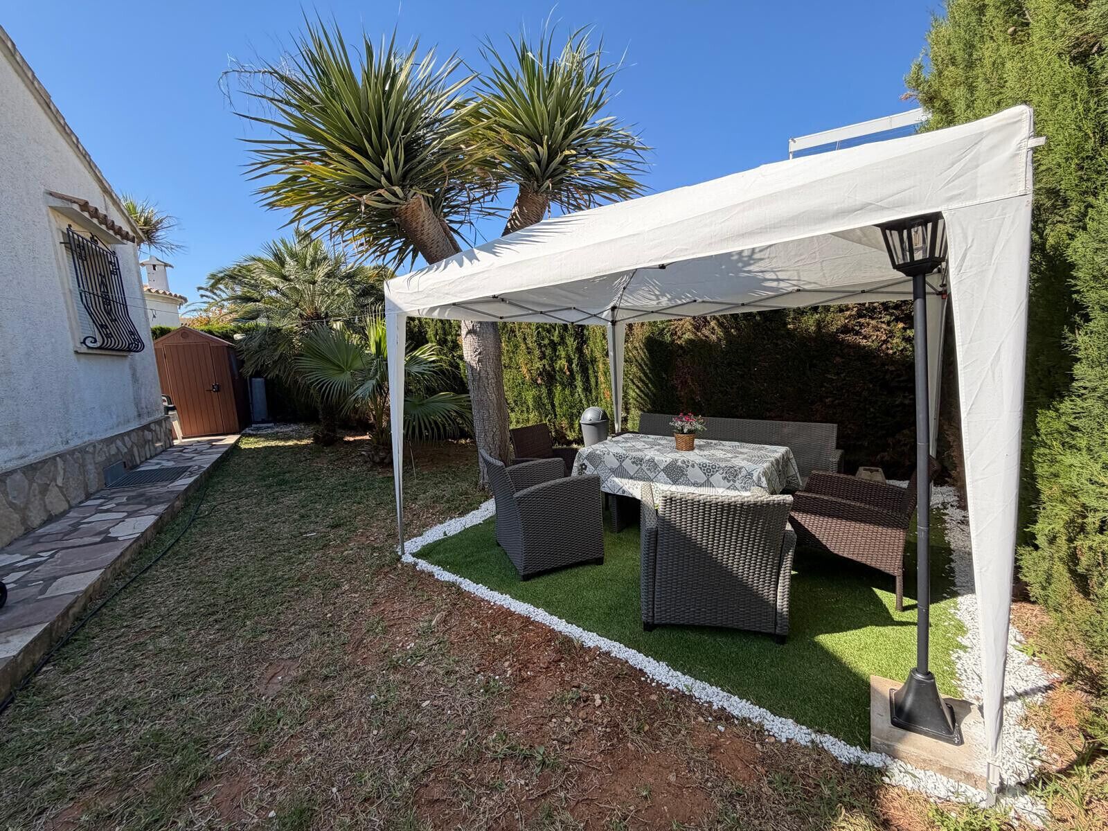 Ein überdachter Patio-Bereich mit Korbmöbeln und einem Tisch, umgeben von einem Garten mit Palmen und einem weißen Gebäude.