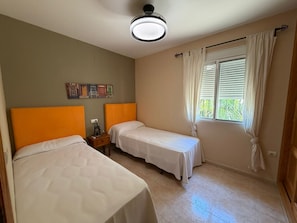 A bedroom with two beds, a ceiling fan, a window with curtains, and a wall-mounted artwork.