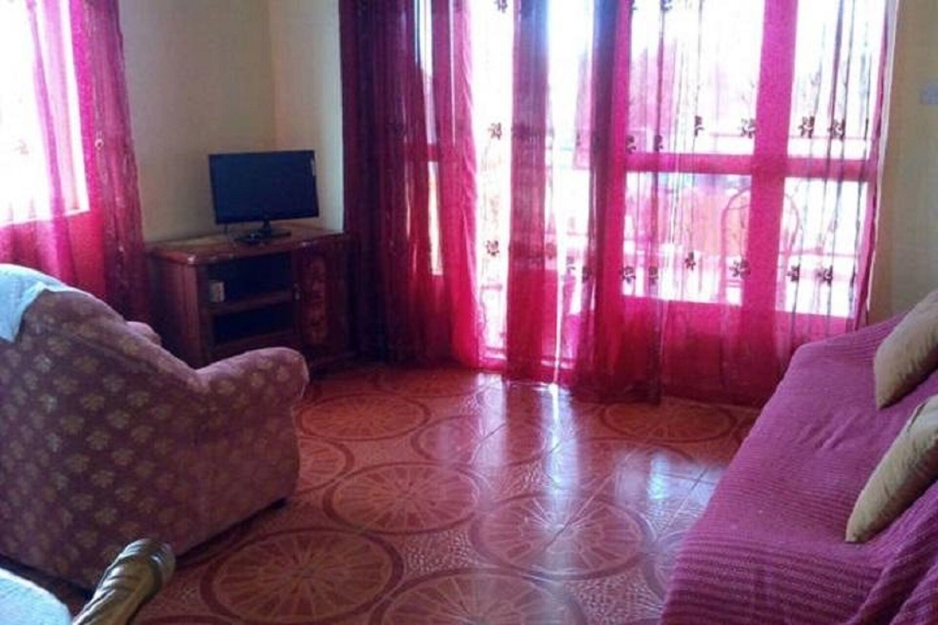 A living room with a patterned floor, a sofa, a television on a stand, and a window with red curtains.