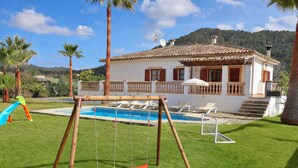 Una casa con piscina, un conjunto de columpios y un patio con sillas de descanso.