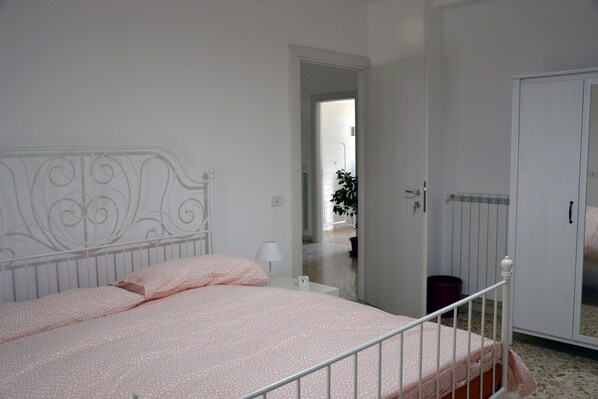 A bedroom with a metal bed frame, a white nightstand with a lamp, and a door leading to another room.