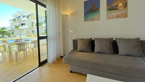A living room with a grey sofa, a dining table with chairs, a large window with curtains, and two framed pictures on the wall. - Sol Nascente- Pool, Cozy, Wifi, Calm area (Vilamoura)