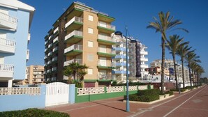 Un immeuble résidentiel à plusieurs étages, doté de balcons, d’une entrée sécurisée et d’un chemin piétonnier dallé bordé de palmiers.