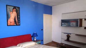 A bedroom with a bed, a red headboard, a nightstand with a lamp, a wall-mounted TV, and a door.