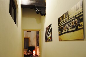 A narrow hallway with a wooden door at the end, a small table with a candle, and two photographs on the wall.