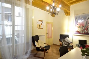 A room with a dining table, two chairs, a black office chair, a chandelier, a window with sheer curtains, and a wall with a geometric painting.