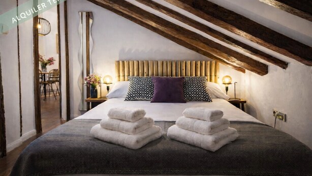 Un dormitorio con techo alto, vigas de madera a la vista, una cama con dos almohadas y una mesita de noche con lámpara.