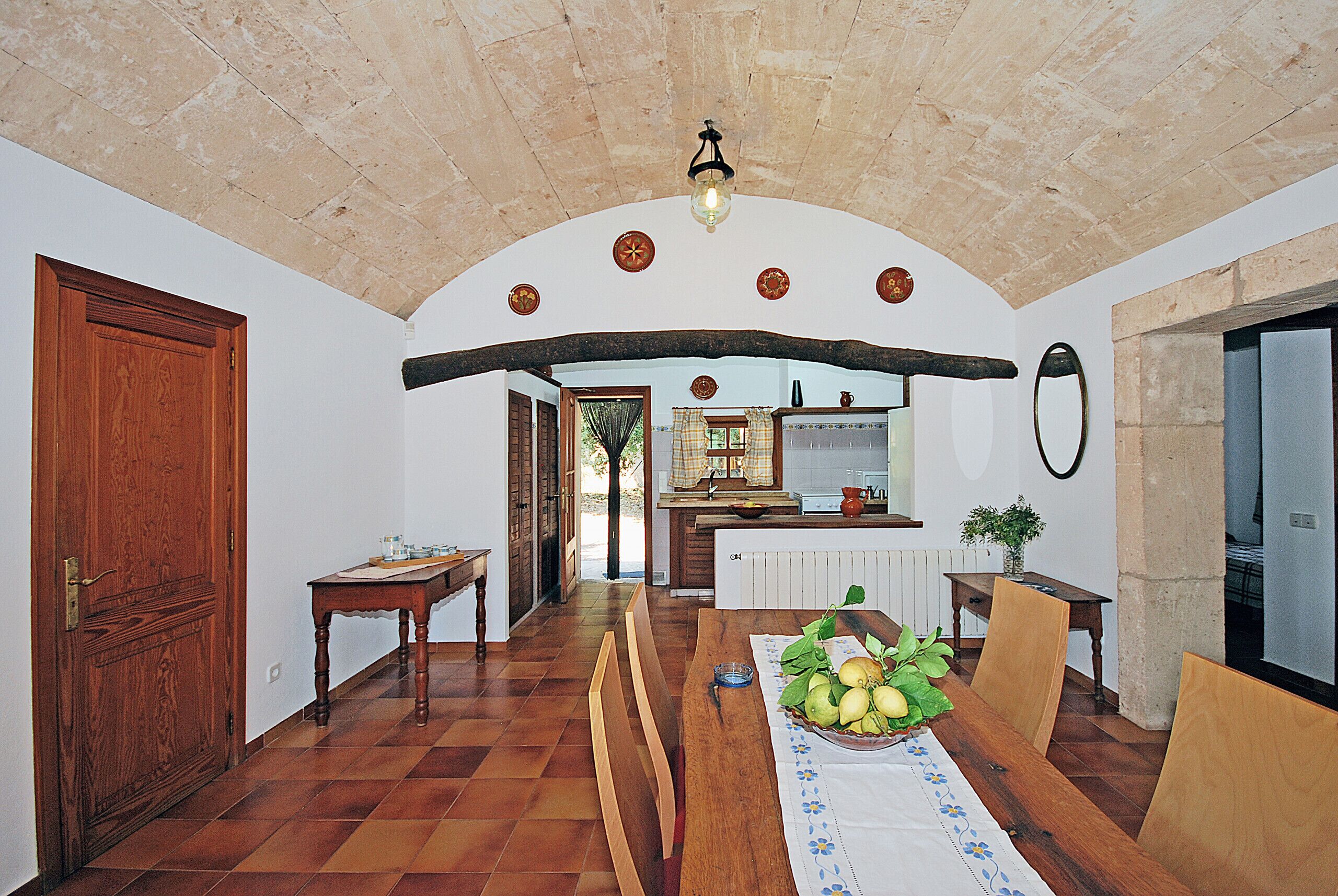 Un coin repas avec une table en bois dressée pour quatre personnes, un saladier rempli de fruits et une vue sur la cuisine.