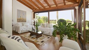 Uma sala de estar ampla com teto de madeira, mobiliário branco, uma mesa de centro de vidro e uma grande janela com vista para o jardim.