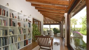 Um quarto espaçoso com teto de madeira, uma estante grande cheia de livros, uma poltrona confortável e vista para um jardim através de janelas amplas.