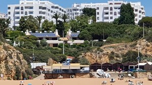 A coastal area with white buildings, a rocky hillside, and a beach with people and parked cars.